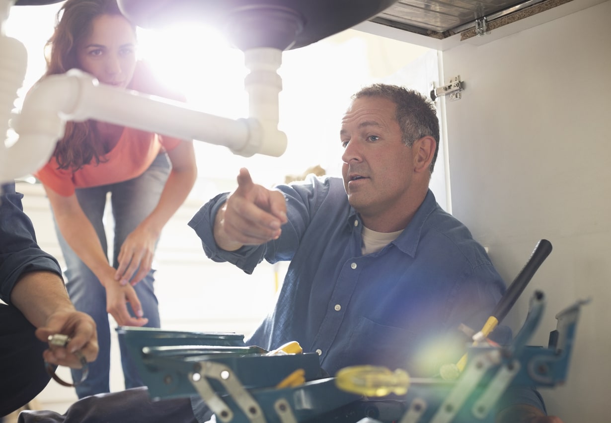 A plumber works on pipes underneath a sink while pointing at them for the client.